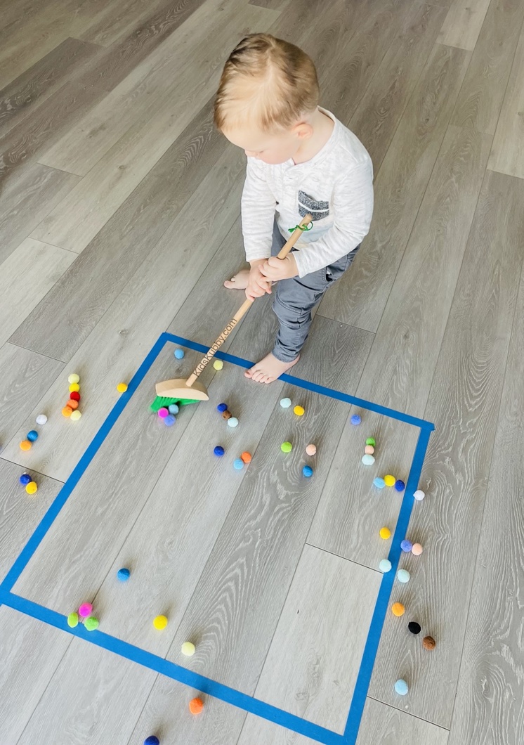 Chore Sweep | A fun and easy activity for toddlers using painters or masking tape! Let them sweep pom poms into shapes you make on the floor with tape. So easy and fun! Plus, your little ones feel like they are involved with the chores. 