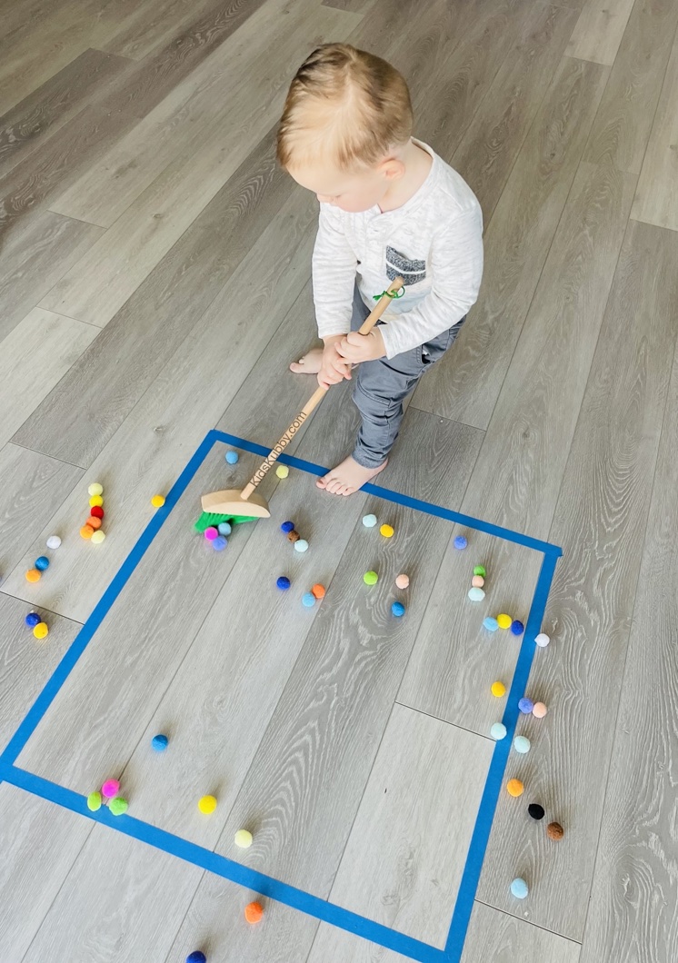 Chore Sweep | A fun and easy activity for toddlers using painters or masking tape! Let them sweep pom poms into shapes you make on the floor with tape. So easy and fun! Plus, your little ones feel like they are involved with the chores.