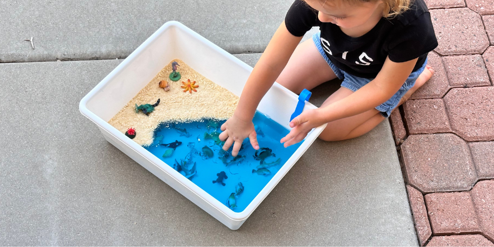 The perfect sensory play idea for toddlers. Let them have sticky messy fun with this easy to make ocean themed sensory bin using Jello! All you need is a container, blue jello, and some small sea creatures. Kids can squish the jello and use tongs to find the sea animals. This is a great way to introduce different senses to babies too! And the sensory bin is taste safe. Perfect for young kids.