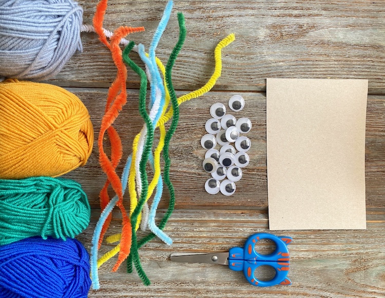 Materials needed for making yarn monsters including yarn, pipe cleaners, googly eyes, scissors, and cardboard. 