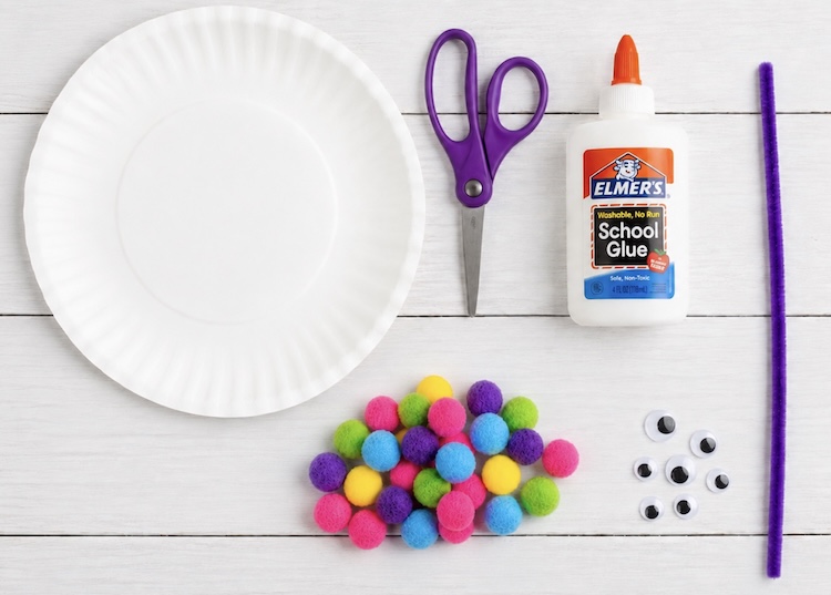 A paper plate, scissors, glue, pom-poms, a craft pipe cleaner, and googly eyes for making craft caterpillars.