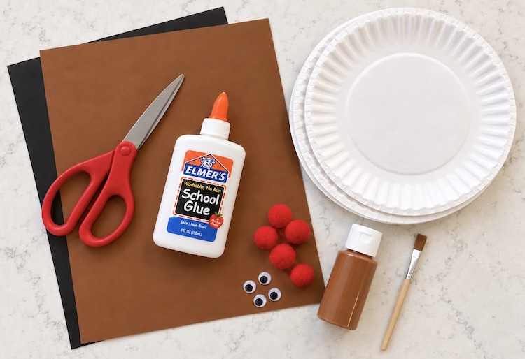 Paper plates, construction paper, glue, scissors, paint, pompoms, and googly eyes all set out on a counter ready for making an easy reindeer craft for Christmas.