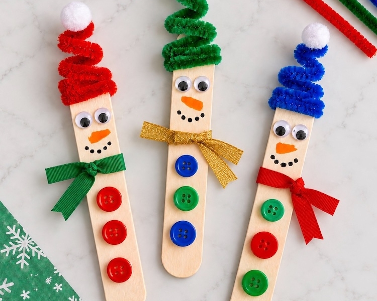 How to make adorable snowmen for Christmas using popsicle sticks and pipe cleaner hats, ribbon scarves, and buttons. A fun and easy craft for kids to make at home. Use them as tree ornaments, magnets, holiday decorations, or trinkets.