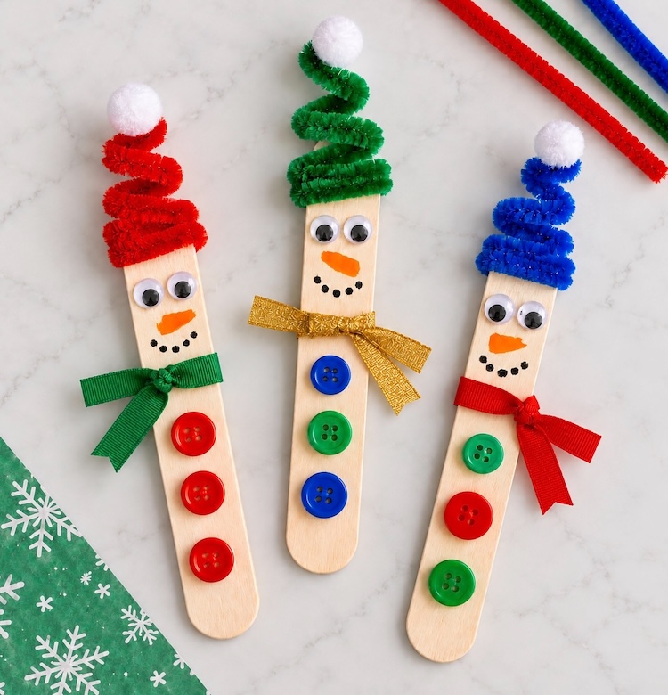 How to make adorable snowmen for Christmas using popsicle sticks and pipe cleaner hats, ribbon scarves, and buttons. A fun and easy craft for kids to make at home. Use them as tree ornaments, magnets, holiday decorations, or trinkets.