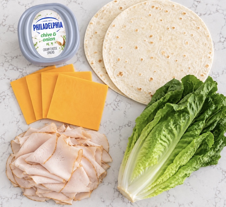 Sliced cheddar cheese, deli sliced turkey, flour tortillas, romaine lettuce, and a seasoned cream cheese spread on a counter.