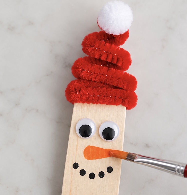 Close up view of a popsicle stick snowman with a red pipe cleaner hat, googly eyes, and a cute carrot nose painted on.