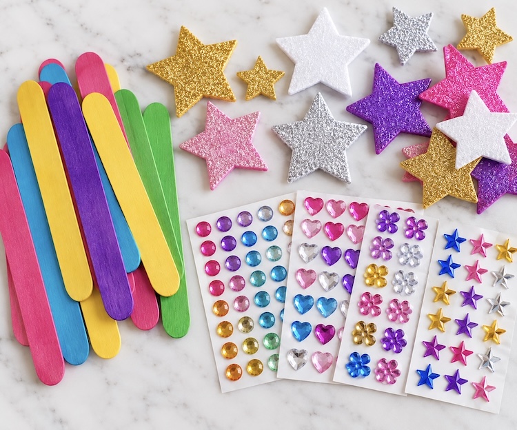 Colorful popsicle sticks, star shaped foam stickers, and small gem stickers on a counter, ready to make magic reading wands for kids.