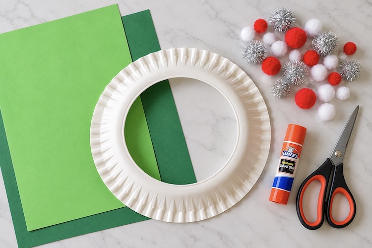 A paper plate, green construction paper, colorful pom poms, scissors, and a glue stick laying on a counter ready to make a Christmas wreath craft.
