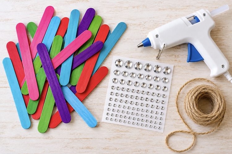 Colorful popsicle sticks, rhinestone stickers, a hot glue gun, and twine for making DIY snowflake Christmas ornaments.