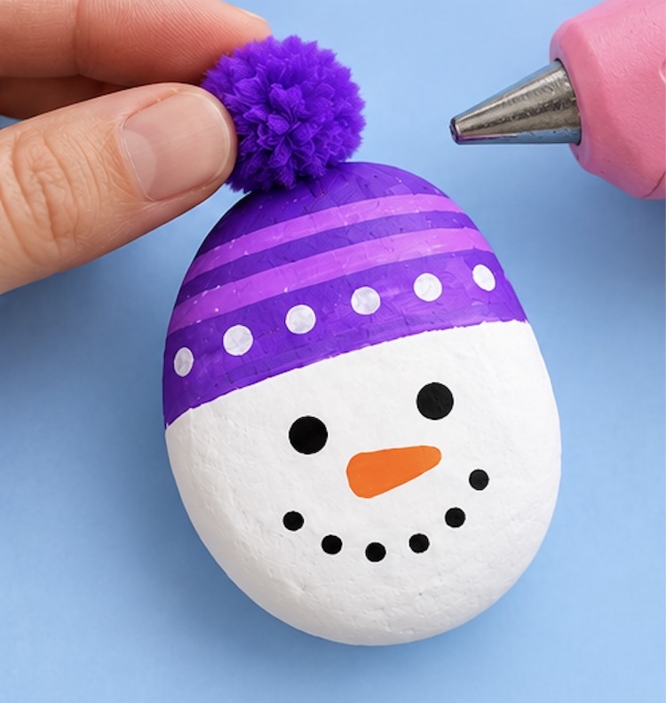 Last step of making a snowman craft with rocks, gluing a Pom Pom onto the top of the winter hat.