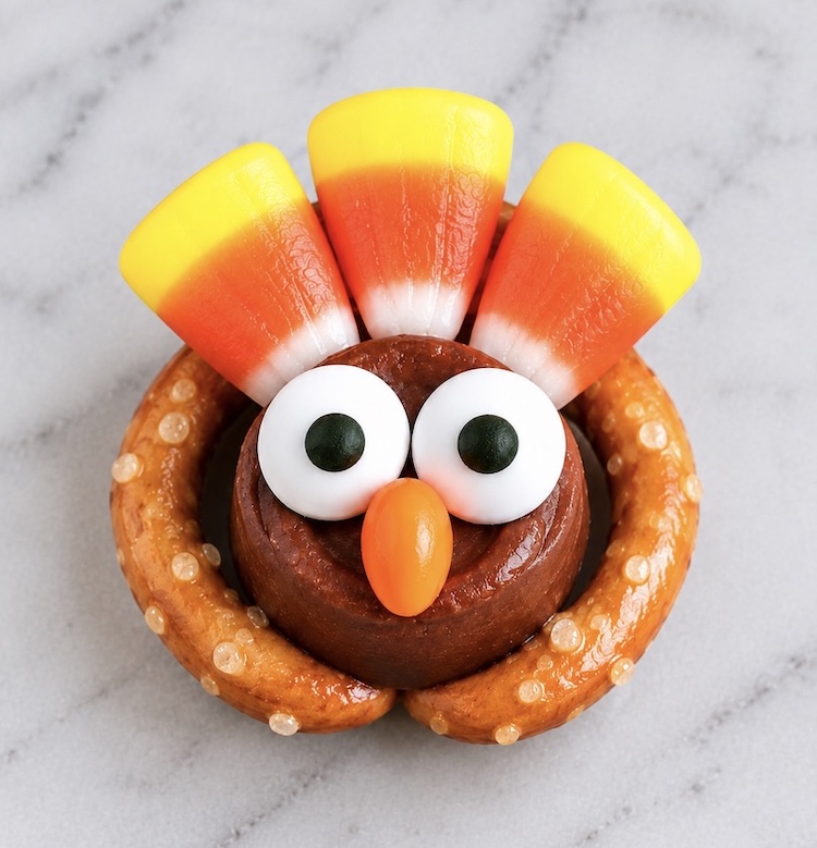 Close up view of a mini chocolate turkey treat made for thanksgiving featuring a pretzel twist topped with a Rolo candy that has been partially melted and then topped with candy to look like a little edible turkey. 