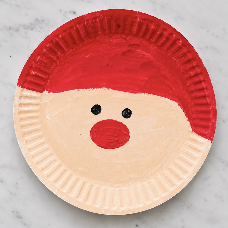 Step two of making a paper plate santa, coloring the plate with paint with a red hat, eyes, red nose, and skin tone. 