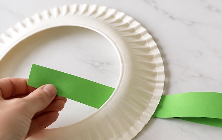 Wrapping green strips of paper around a paper plate that has been cut into the shape of a wreath.