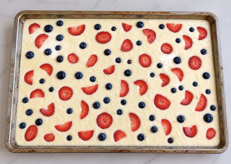 Step two of making sheet pan pancakes in the oven. Top the batter with some optional toppings such as berries, bananas, chocolate chips, nuts, etc.