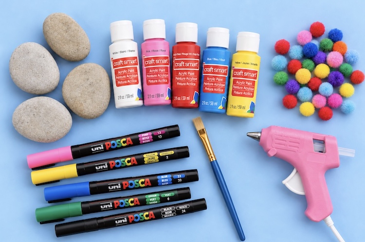 Craft supplies needed to make cute snowman rocks including flat rocks, acrylic paint, paint pens, pom-poms, and a hot glue gun.